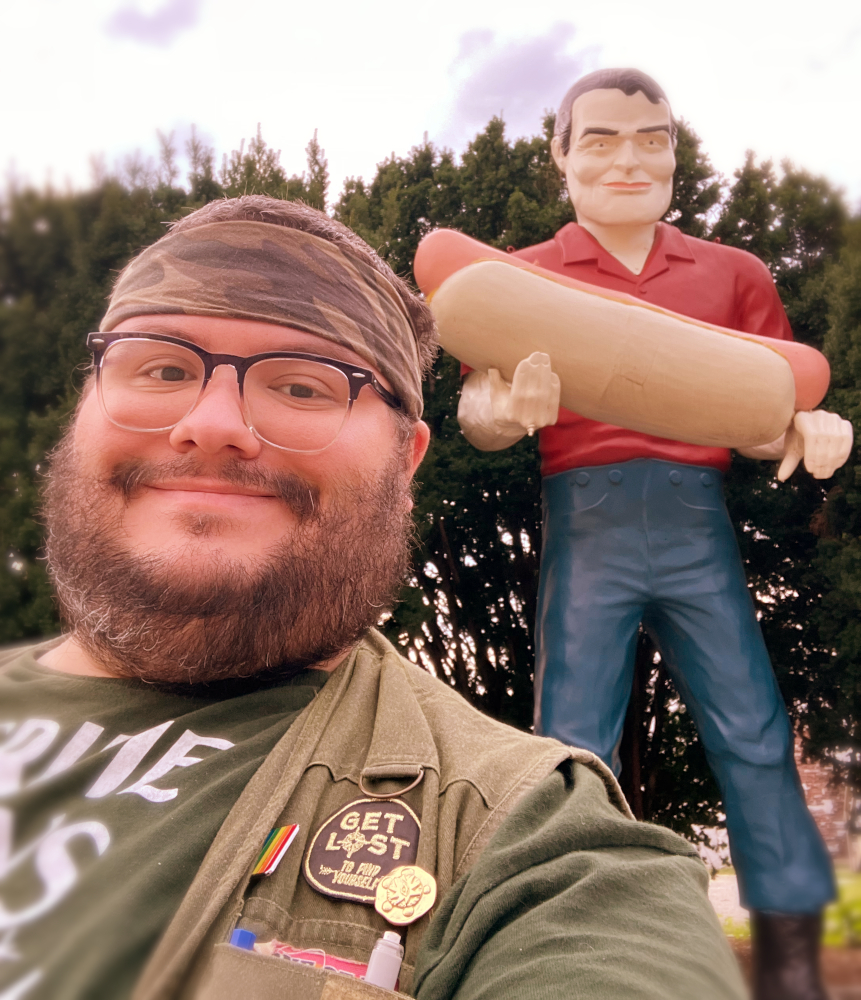 The author of Puzzle Road Trip: Route 66, Dave Millar, in dark green shirt, vest, and bandana, taking a selfie in front of a giant statue of a clean-cut Muffler Man statue with the man holding a hot dog.